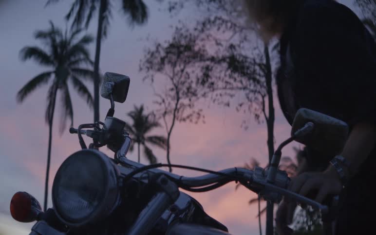 A long-haired biker gets on his chopper and watches the tropical sunset behind silhouetted palm trees