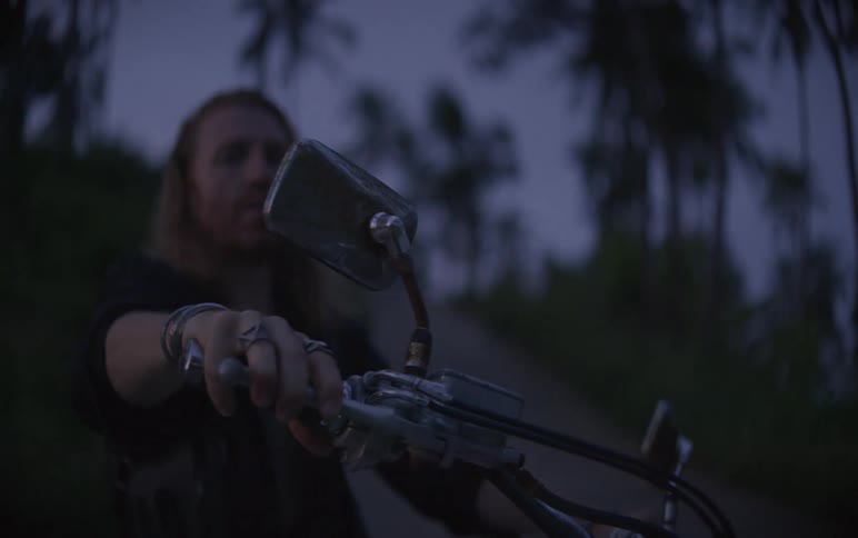 A long-haired biker gets on his chopper motorcycle at dusk on a quiet, tree-lined road