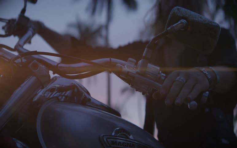A long-haired biker gets on a motorcycle at dusk, turning on the headlight with palm trees silhouetted against a colorful sunset sky