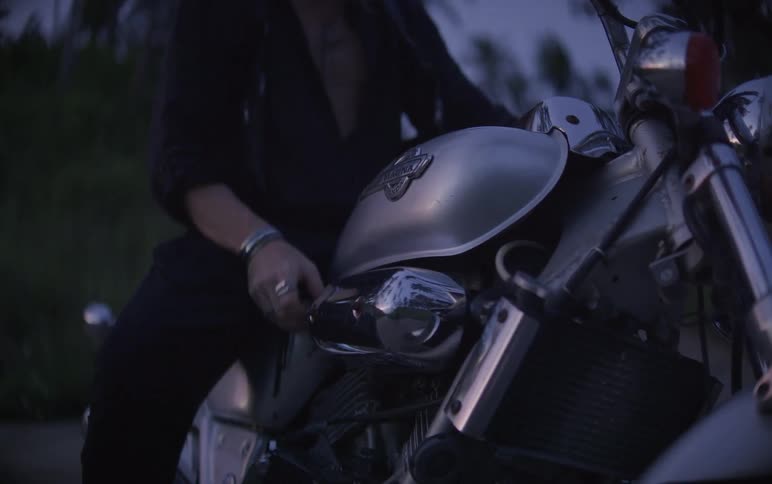 A man in black mounts his silver motorcycle at dusk, puts on his gloves, and turns on the headlight