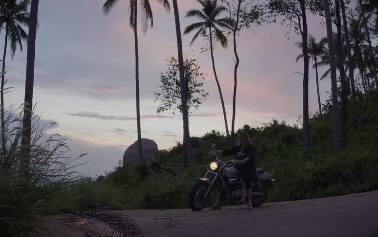A woman on a motorcycle pulls over on a lush, tropical road to enjoy the beautiful pastel sunset sky