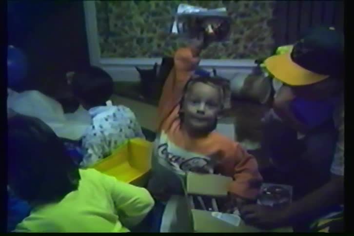 A happy little boy opens a gift and triumphantly holds up his new toy at his birthday party in this nostalgic home video