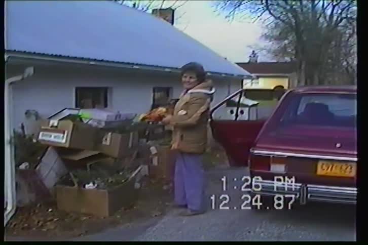 A smiling woman holding a fruit basket waves to the camera outside a garage on Christmas Eve in 1987