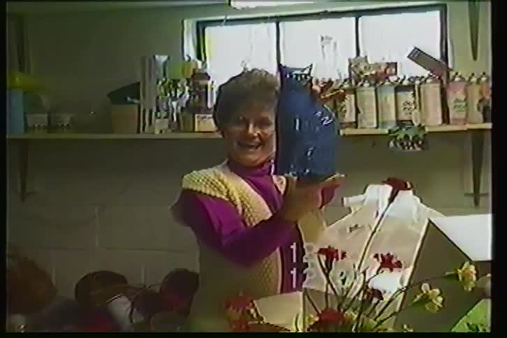 A smiling woman in a flower shop proudly displays a unique blue cat-shaped vase she received as a gift in 1987