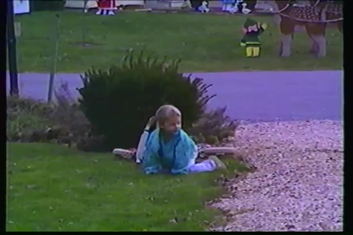A little girl on a scooter falls onto the grass and smiles as her sibling rides by on another scooter during Christmas time