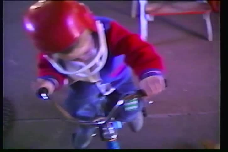 A young boy wearing a red and white football helmet slowly rides his bicycle inside a garage