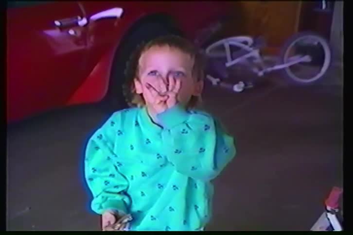 A funny young boy with a mullet and blue eyes gives a thumbs-up in a garage in a vintage home video
