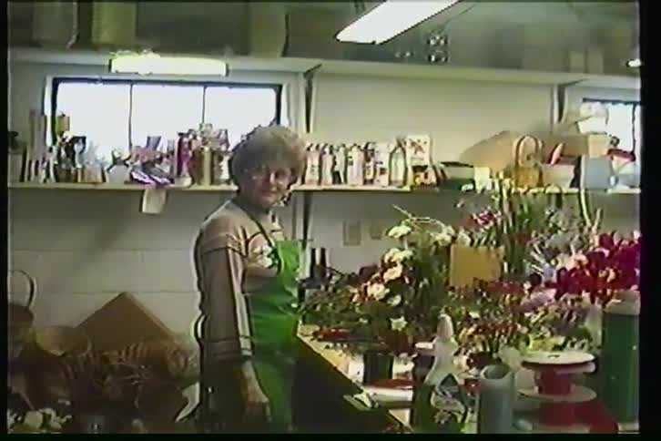 A woman in a green apron smiles and waves from her workshop before trimming evergreen branches in a vintage home video