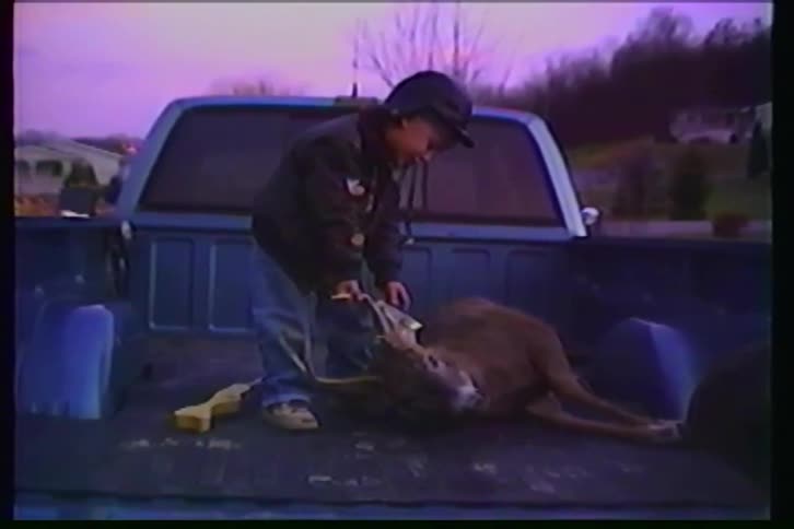 A young boy proudly tags a hunted deer in the back of a pickup truck at dusk