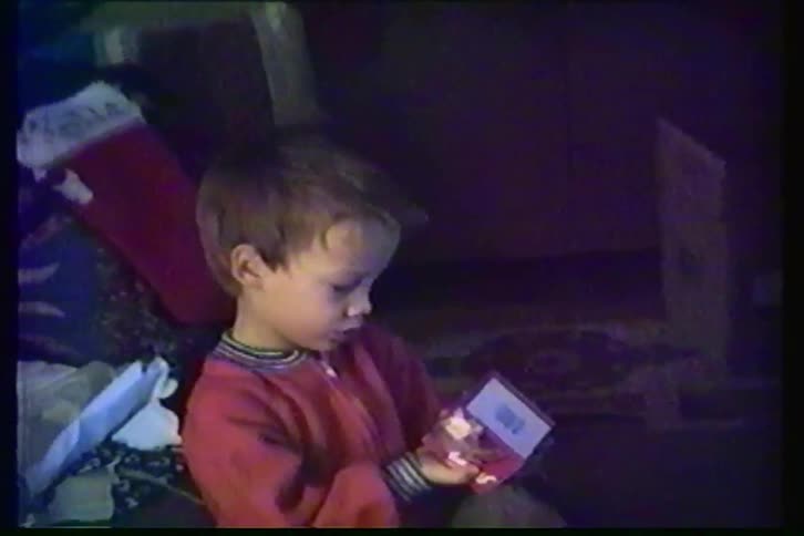 A nostalgic home video from the 1990s shows a young boy in red pajamas excitedly inspecting a small Christmas gift