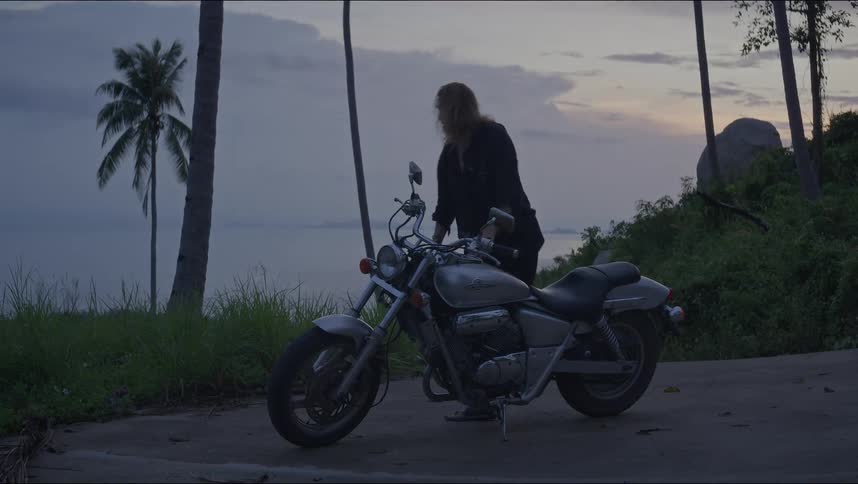 A man with long hair gets off his motorcycle parked by the ocean at dusk and walks away