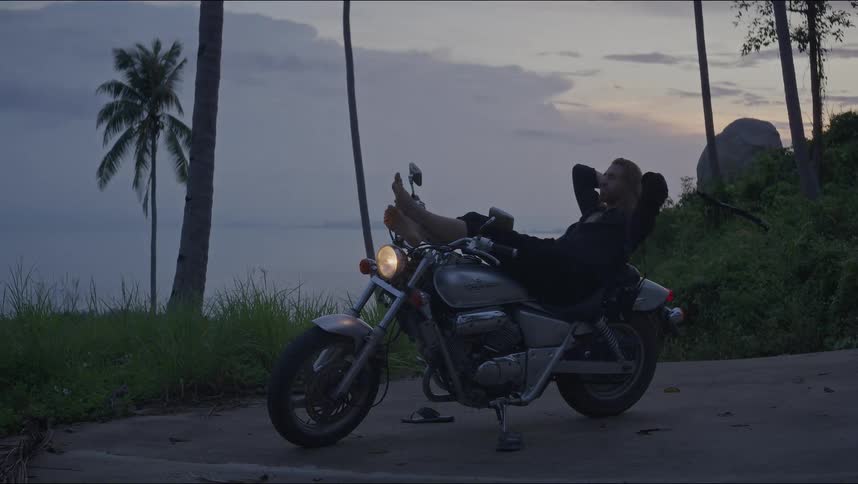 A man with long hair relaxes on his motorcycle, enjoying the tranquil tropical ocean view at dusk
