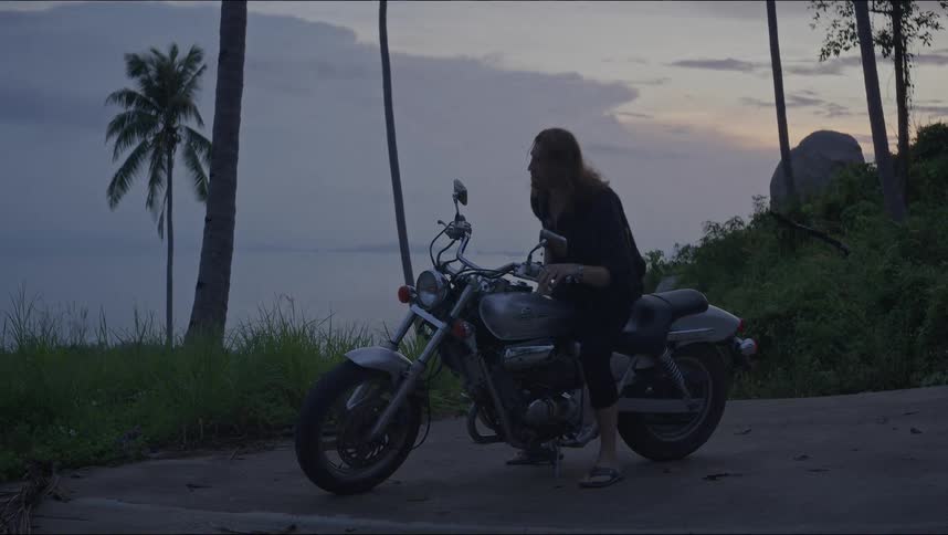 Long-haired biker parks their motorcycle on a scenic tropical road by the sea at sunset and walks away