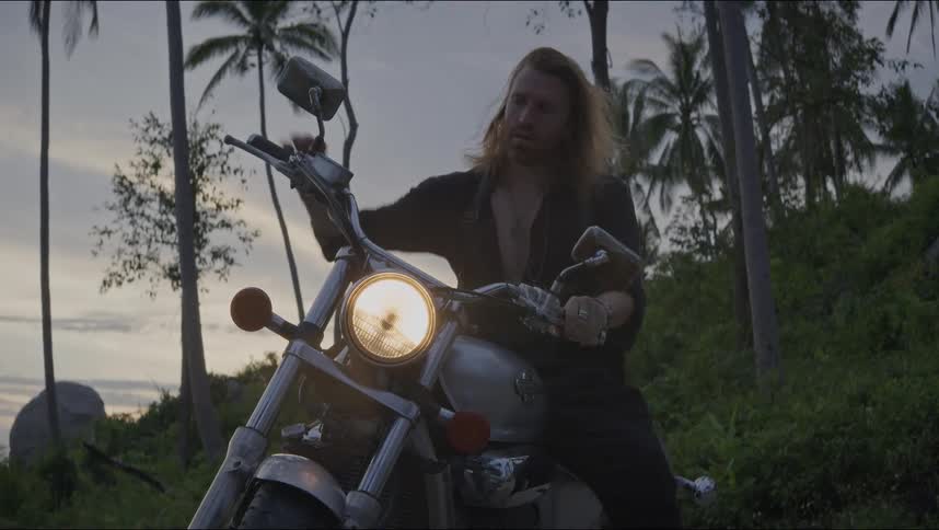 A rugged, long-haired man gets on his chopper motorcycle and starts the engine in a tropical jungle at dusk