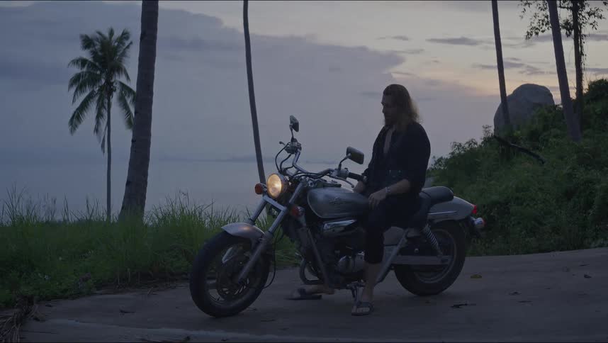 A long-haired man relaxes on his motorcycle while enjoying the scenic ocean view from a tropical island road at dusk