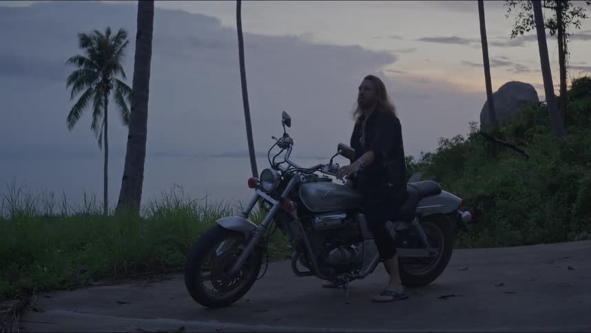 A long-haired man gets on his motorcycle parked on a tropical coastal road at dusk before starting his journey