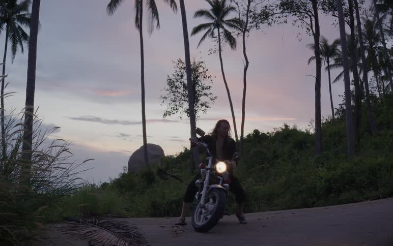 A biker with long hair turns on his motorcycle and rides away down a tropical road at dusk