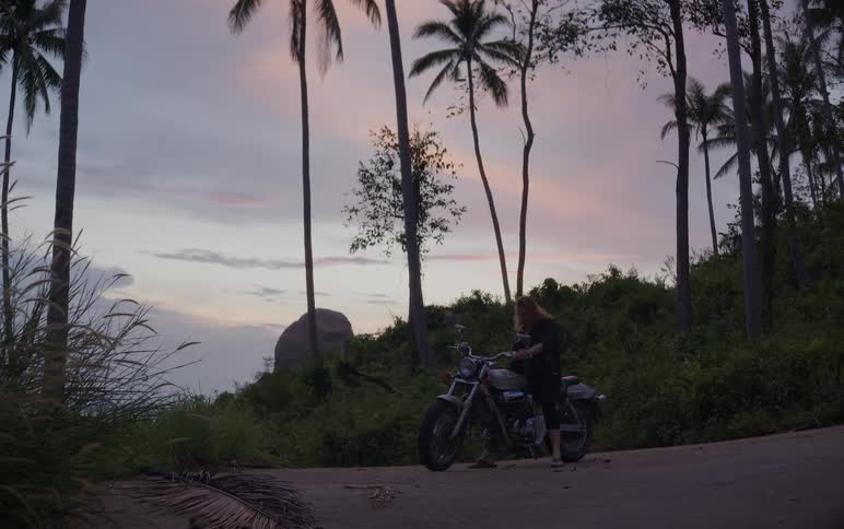 A long-haired person walks to a motorcycle parked on a rural tropical road, gets on, and turns on the headlight against a beautiful sunset sky