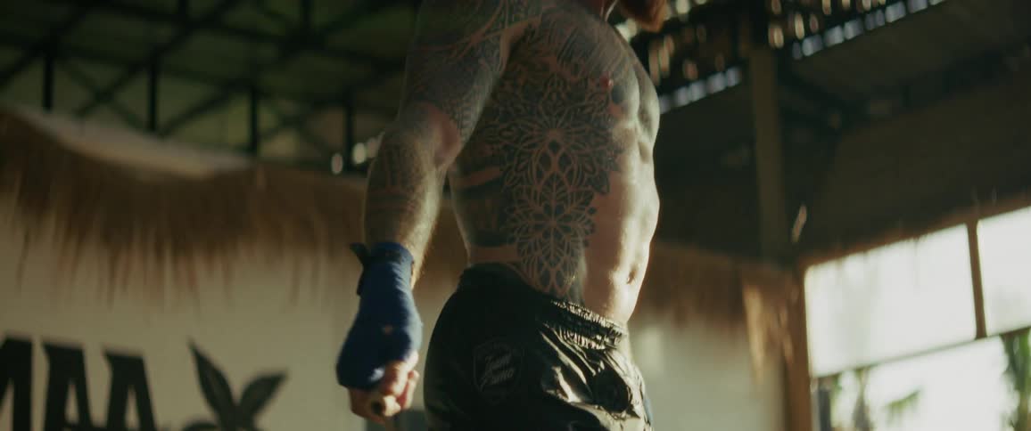 A close-up slow-motion shot pans up from the tattooed feet to the face of a muscular athlete as he jumps rope barefoot in a gym