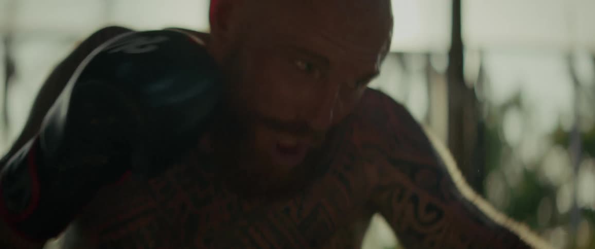 A close-up slow-motion shot shows a muscular, tattooed boxer intensely training in a sun-drenched gym, with sweat flying from his body as he punches