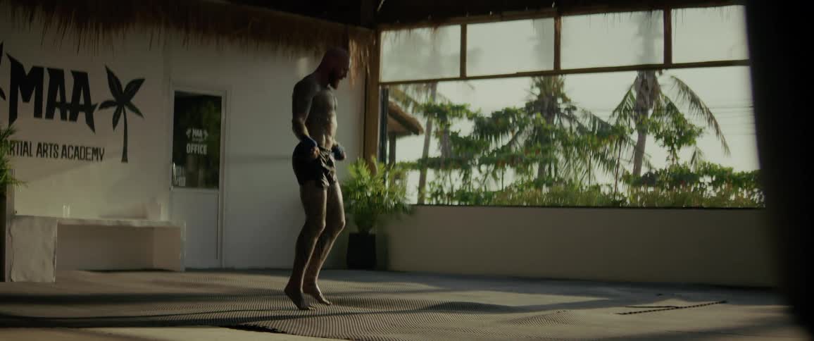 Slow motion shot of a bald, tattooed fighter warming up with a jump rope in a gym with a view of palm trees