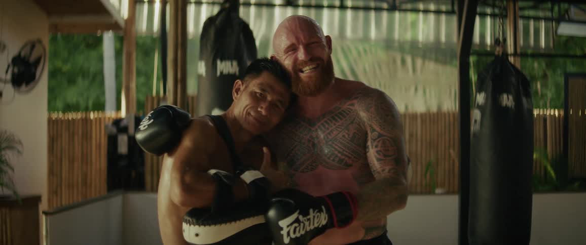 A sweaty, tattooed Muay Thai fighter and his trainer smile and pose happily together after a training session in a tropical gym