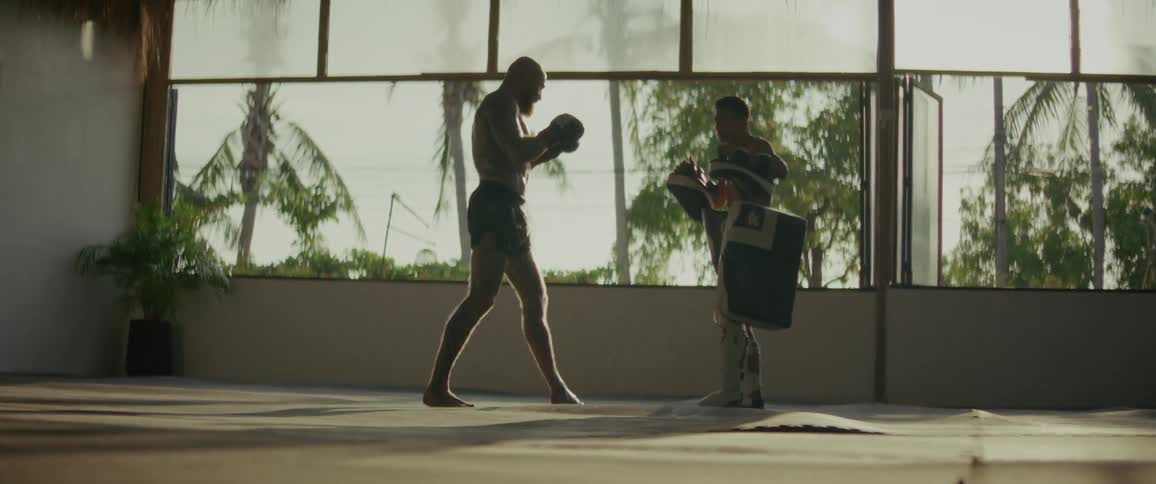 A tattooed fighter and his trainer practice powerful punches and kicks in slow motion in a tropical open-air gym