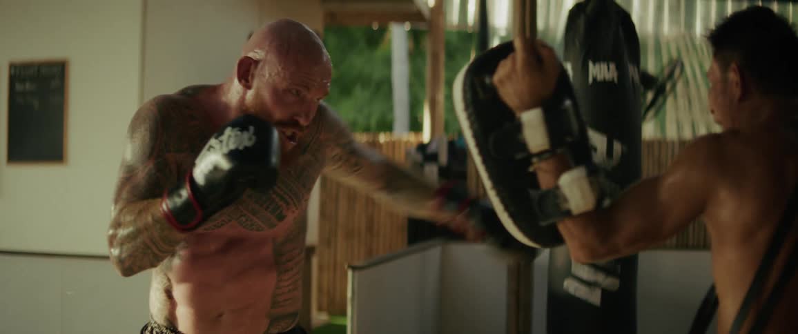 A heavily tattooed fighter intensely trains with his coach, throwing powerful punches and kicks in slow motion