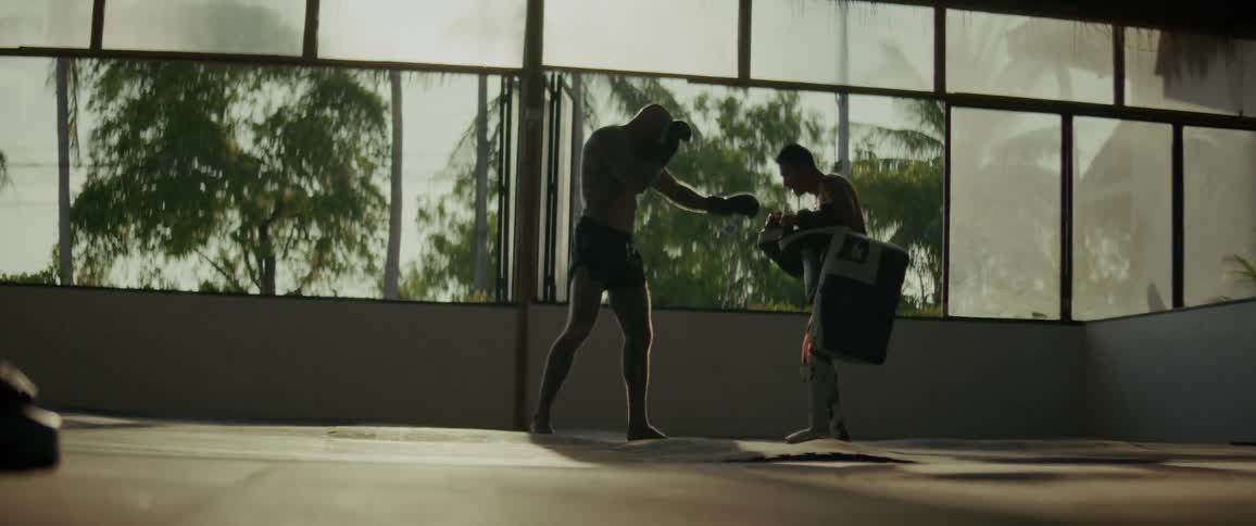 Two men practice Muay Thai in slow motion, silhouetted in a gym with a tropical background