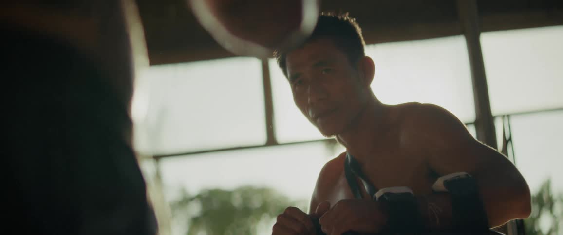 An intense, close-up slow-motion shot of a Muay Thai coach holding focus pads for a fighter during a training session with dramatic sun flare