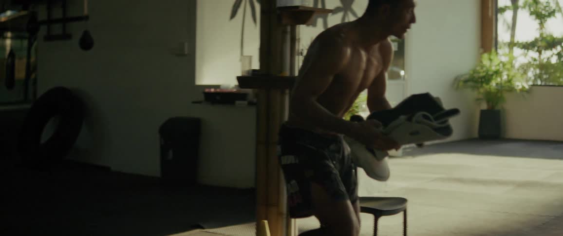 A muscular fighter walks from the shadows into a sunlit martial arts studio and prepares for training by putting on his shin guards in slow motion