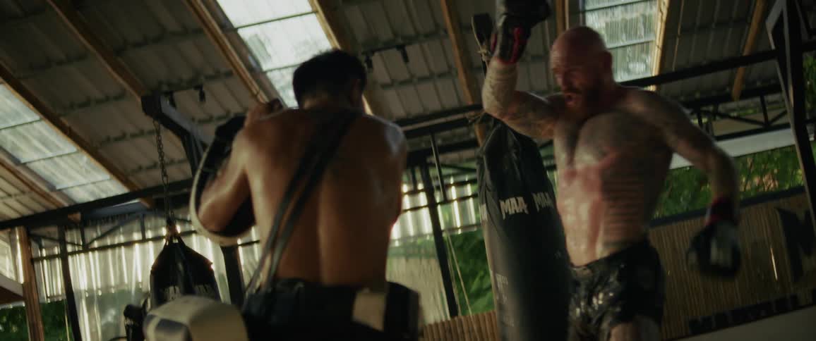 A muscular, tattooed fighter practices powerful Muay Thai kicks and punches with his trainer in a gym during a slow-motion training session