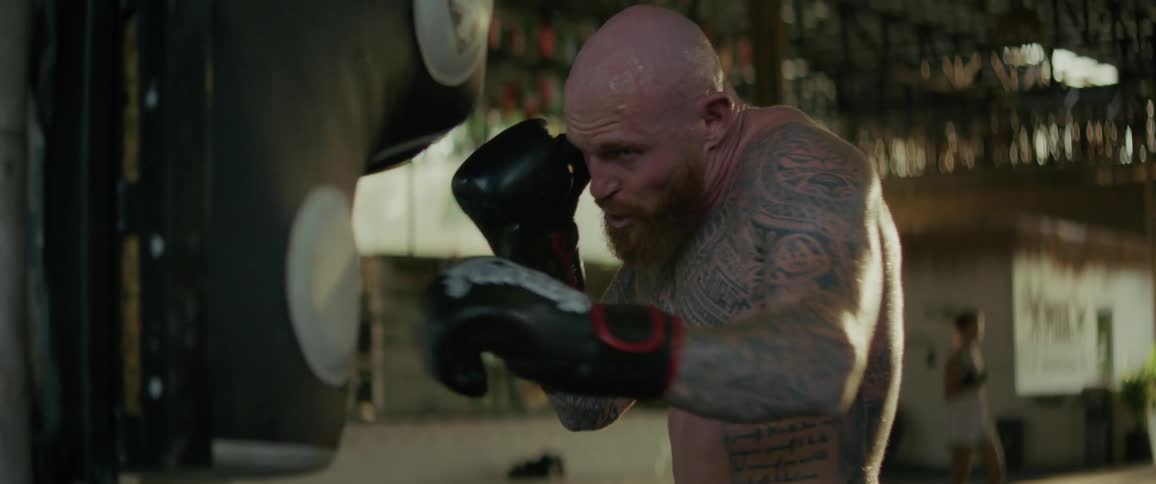 A powerful, tattooed boxer intensely trains on a punching bag in a gritty gym in slow motion