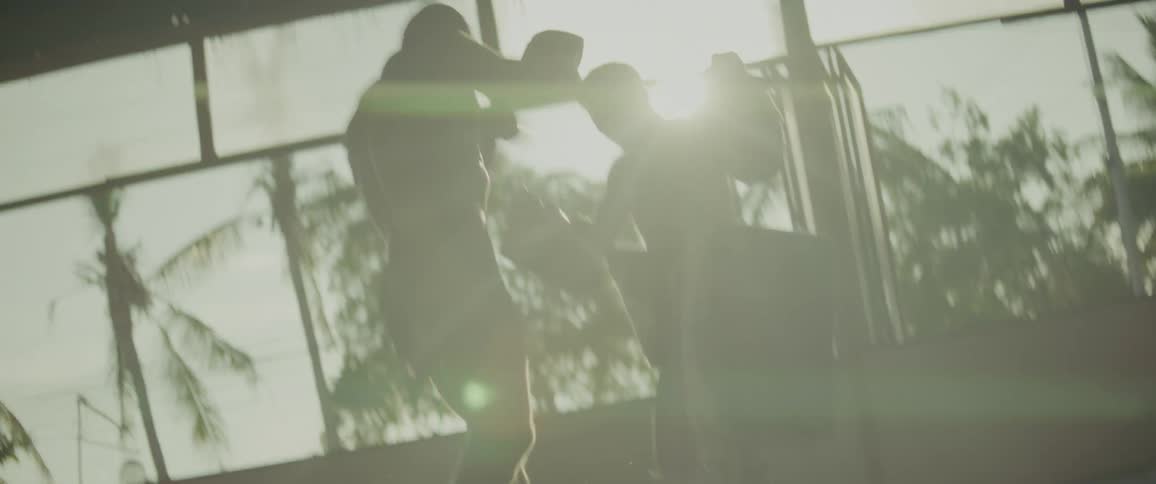 A tattooed Muay Thai fighter practices powerful kicks and punches with his trainer in a sun-drenched, open-air gym