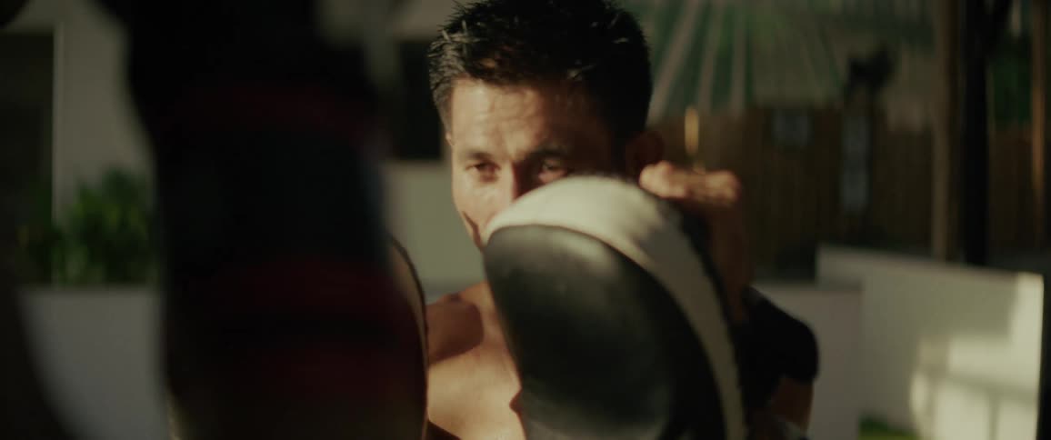 Intense close-up of a focused Muay Thai trainer holding pads for a student in slow motion during a sunlit workout