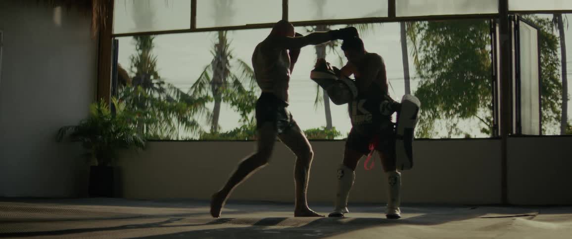 A powerful tattooed fighter and his trainer practice punches and kicks in slow motion, silhouetted against a tropical gym window