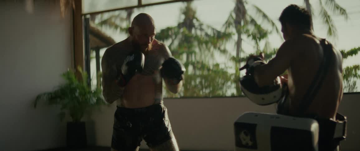 An intense, tattooed fighter practices powerful punches and kicks with his trainer in slow motion against a sunlit tropical backdrop