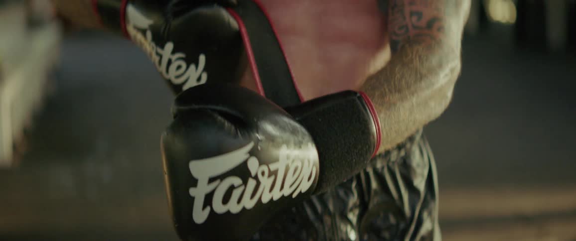 A tattooed fighter stands prepared with his black boxing gloves on in a gym, filmed in slow motion