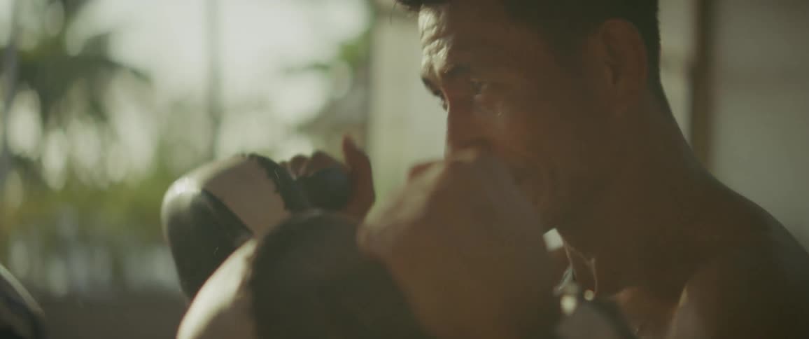 An intense boxer in slow motion relentlessly strikes a punching pad held by his trainer in a hazy, sunlit gym