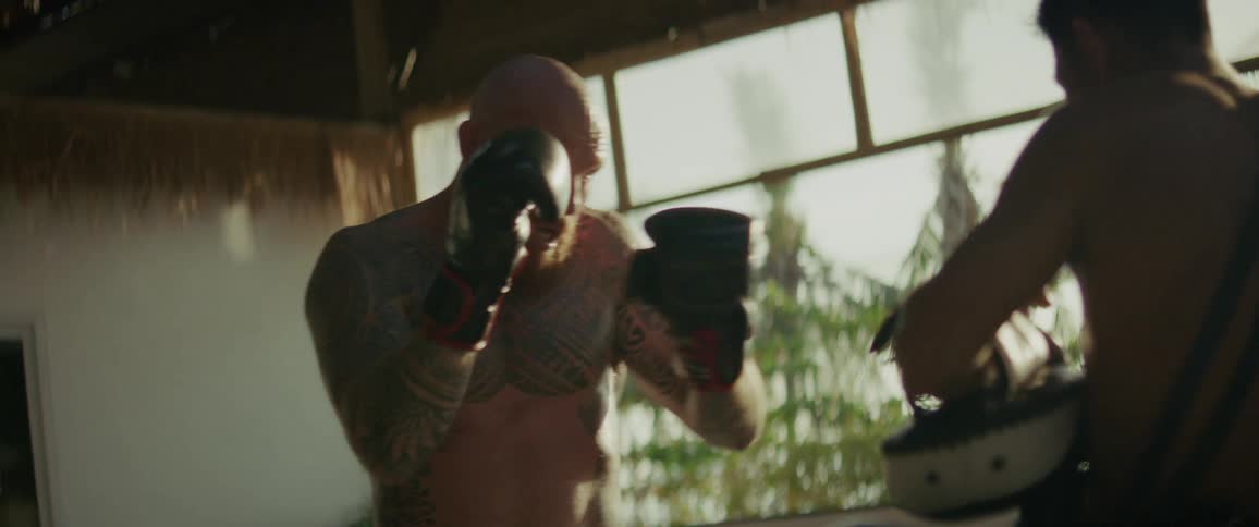 A powerful, tattooed fighter practices kickboxing with his trainer in a tropical gym in slow motion