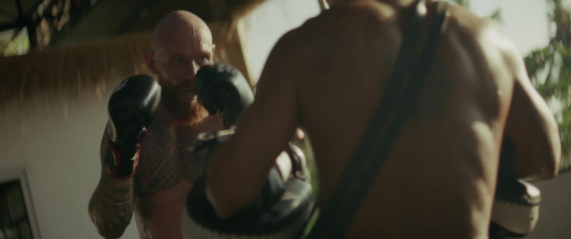 In slow motion, a powerful, tattooed fighter relentlessly trains with his coach, throwing punches and kicks in a sunlit tropical gym