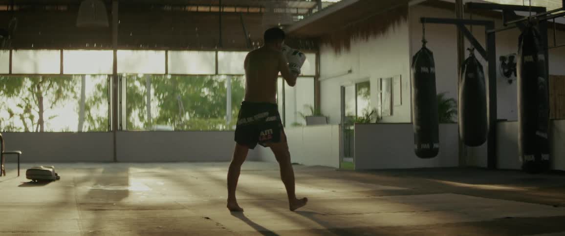 A Muay Thai fighter shadowboxes in a sun-drenched gym, practicing kicks and punches in slow motion