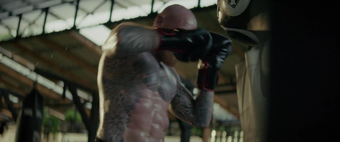 A muscular, tattooed boxer relentlessly punches a heavy bag during an intense training session in a rustic gym
