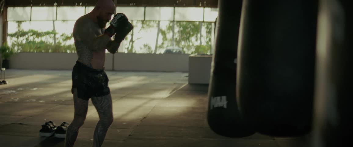 A powerful tattooed fighter practices his punches and kicks on a heavy bag in a sunlit gym in slow motion