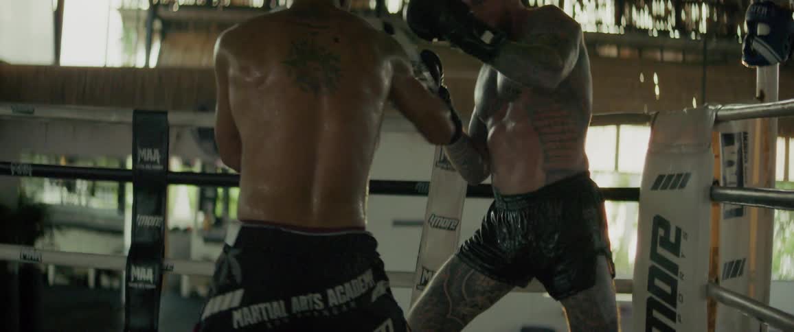 Two powerful Muay Thai fighters with tattoos trade blows and kicks during an intense sparring session in a gritty, open-air boxing ring