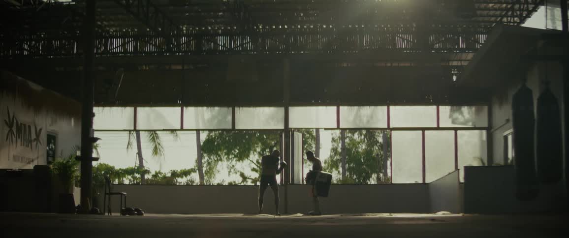Two men are silhouetted while practicing powerful Muay Thai kicks and punches in a sunlit, open-air gym