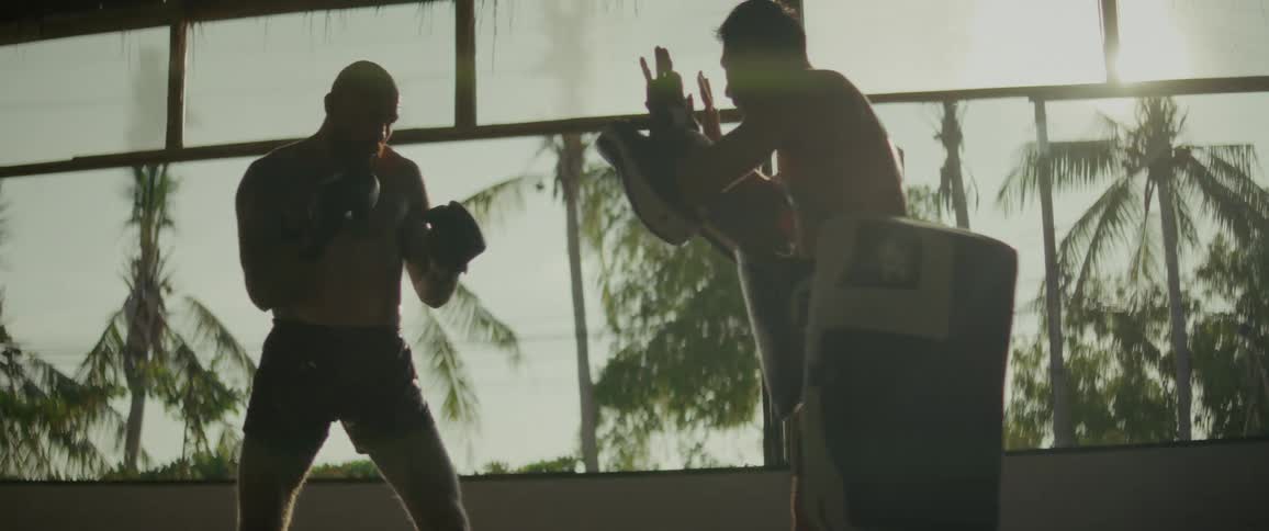 A tattooed fighter powerfully trains with his coach, throwing punches and kicks in dramatic slow motion, silhouetted against the bright sun in a tropical gym