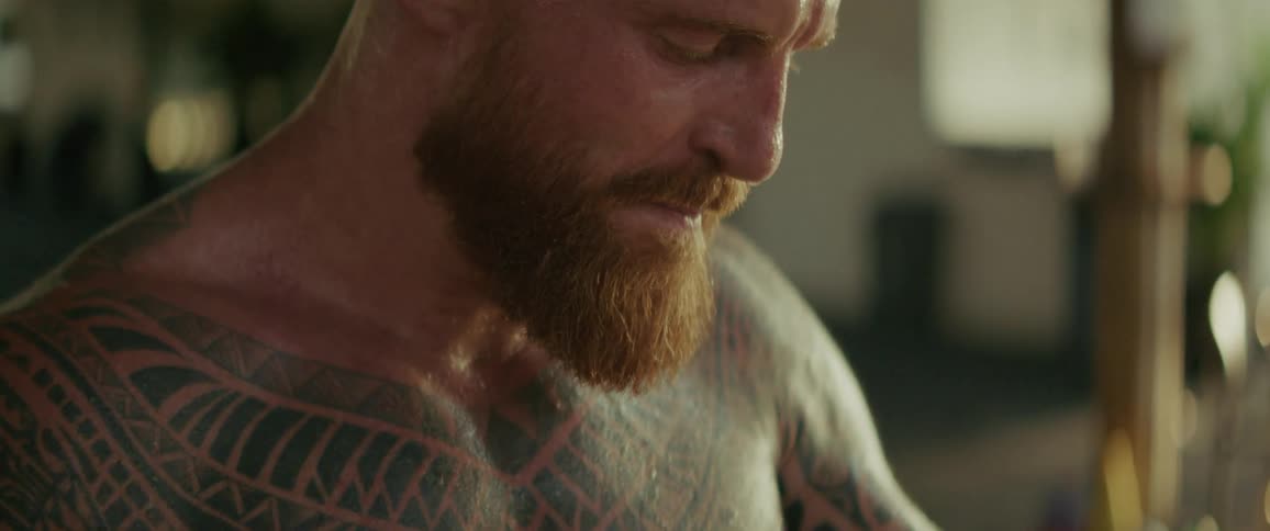 A heavily tattooed boxer with a red beard puts on his boxing gloves with his teeth in a slow-motion close-up shot