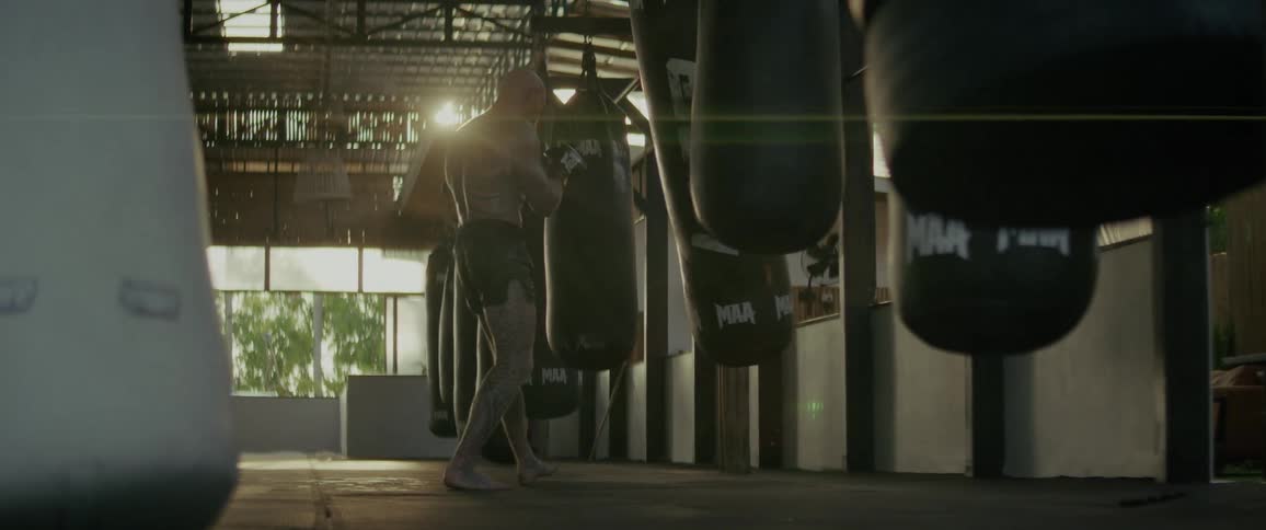 A powerful tattooed boxer punches and kicks a heavy bag in a sunlit gym in slow motion
