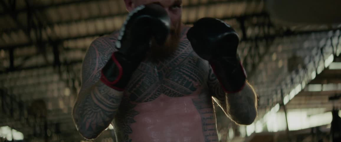 Slow motion low-angle shot of an intense, tattooed boxer with a beard training with a punching bag in a gritty gym